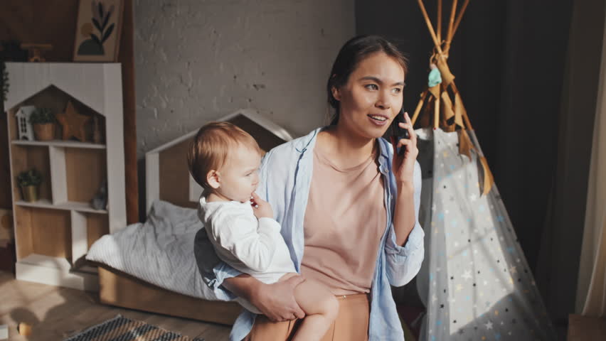 Medium shot of young woman standing in sunlit nursery by window, holding her baby and talking by mobile phone