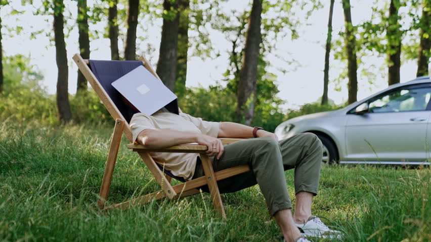 Freelancer sleeps in a folding chair with face covered with laptop. Tired man having a break from work in the nature.