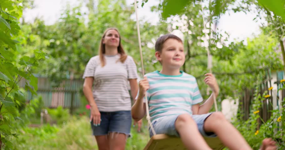 Happy family, mother and child play on wooden swing in garden under tree