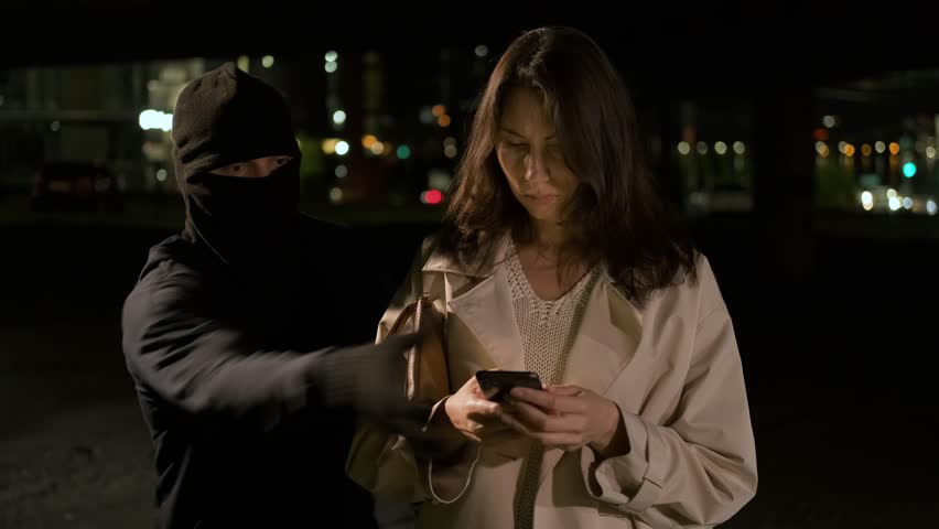 A woman stands at a bus stop with a handbag uses the phone. A masked robber sneaks up from behind, snatches a smartphone and runs away