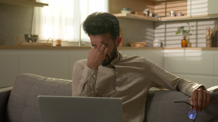 Indian Arabian man exhausted guy freelancer on couch at home kitchen working with laptop overworked suffer eyestrain eyes pain discomfort eye tension take off glasses take break bad eyesight headache Royalty-Free Stock Footage #1105603369