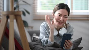 Cheerful young woman waving hand, making video call on smart phone while lying on couch at home.