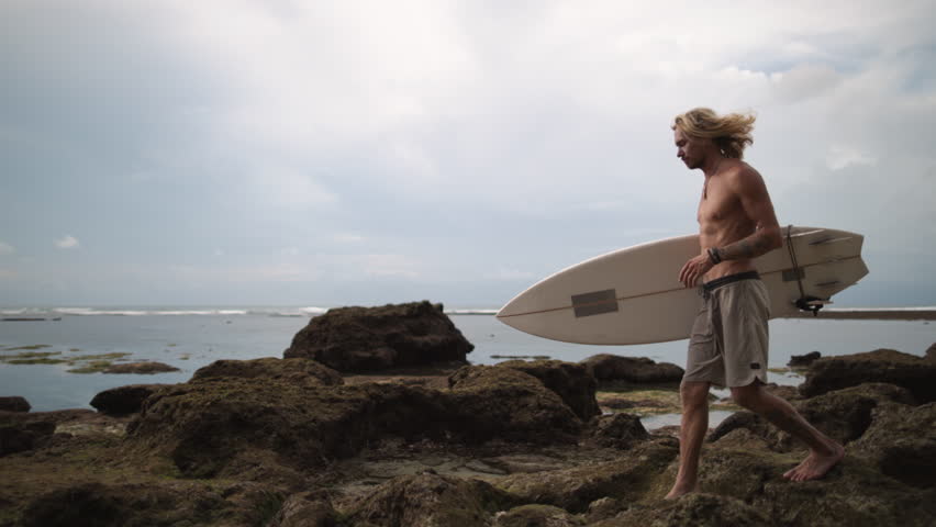 Sporty long-haired Caucasian male surfer walks along coast. Concept of self-development, self-improvement, development of strengths, gaining new experience, increasing confidence and self-esteem.