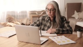 Focused gray haired senior woman studying online at home Curious mature female make notes while talk with teacher by video call on laptop computer Distance remote education for person on pension