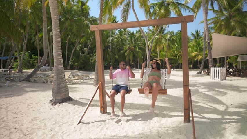 Slow motion of latin american couple playing on a wooden swing between palm trees on a empty tropical beach. Amazing honeymoon on the Caribbean