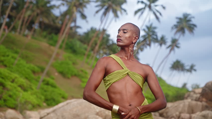 Queer black person in open dress, brass jewelry poses on stone at scenic rocky natural pool by ocean. Lgbtq biethnic gay model in sexy outfit, brass accessories poses on rock. Tropical location.