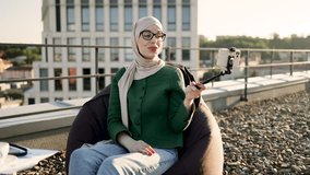 Attractive arabian woman with smartphone on selfie stick filming video content for vlog on roof terrace. Smiling lady in traditional muslim clothing covering topic of work-life balance for followers.