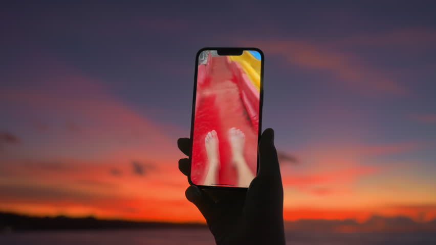 POV watching action camera video from aqua park on a beach at sunset. Summertime. Orange purple dusk and handphone with memories of happy vacation. Cell phone video with feet on a slider at water park
