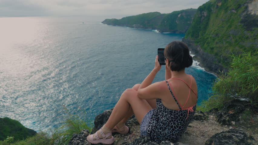 Asian girl relaxing on a rock above sea in summer and recording video on cell phone. Japanese woman holding hand phone sitting on stone at edge of cliff on ocean. Female online social media