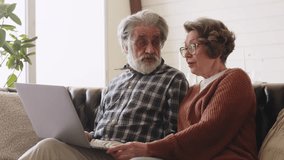 Older couple using computer laptop on sofa at home for online shopping, surfing the web