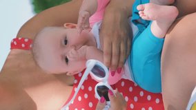 Vertical video. Young mother in swimsuit trying on sunglasses for her cute baby on tropical beach, slow motion. Family vacation and travel concept