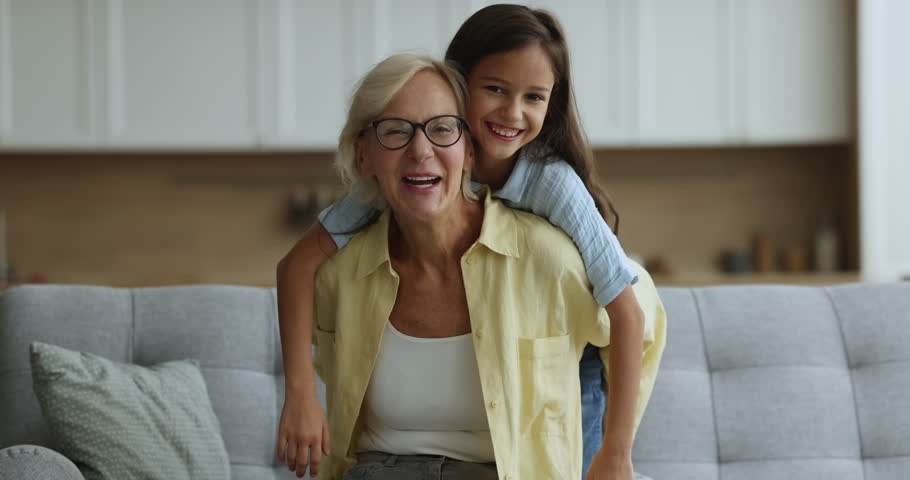 Joyful elderly 60s granny carrying on back little grandchild, girl spread her arms imagines flying like plane, laughing, look at camera, having fun seated on couch at home. Family vacation and travel Royalty-Free Stock Footage #1105653559