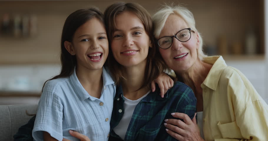 Close up of cheerful older 60s woman her young adult 35s daughter and pre-teen 10s girl, cuddling seated on couch smile look at camera, spend carefree happy weekend together at home, enjoy family ties Royalty-Free Stock Footage #1105653569