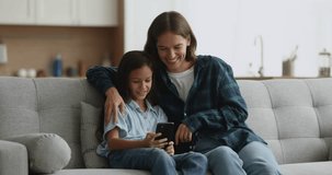 Beautiful young mother hug little daughter sitting on couch at home, enjoy weekend with modern smart phone looking at screen laugh, watch social media video, play online game, use modern application