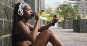 Animation of cloud, arrow icons, african american woman watching video over phone and drinking water. Digital composite, exercise, fitness, cloud computing, technology, futuristic, thirsty, sitting.