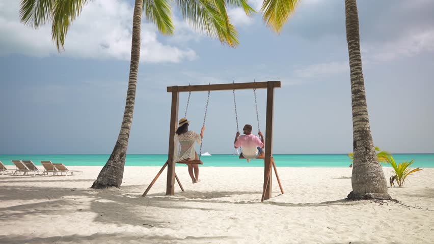 Back view of latin american couple playing on a swing between palm trees on a empty tropical beach. Turquoise water and white sand. Amazing honeymoon on the Caribbean