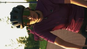 Professional female cyclist riding bicycle on cycle path in park. Vertical video. Young fit confident stylish woman athlete in black helmet and glasses cycling at sunset, doing cycling training