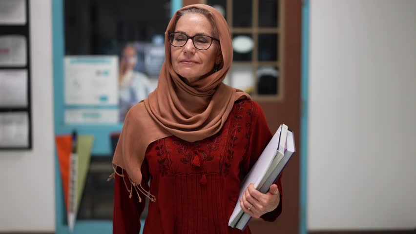 Slow motion front view closeup of happy smiling muslim teacher wearing headscarf walking down a hallway in an empty school holding books.