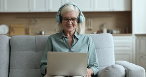 Cheerful positive elder blonde lady in trendy headphones waving friendly greeting hand hello at laptop, speaking on online video conference call, using hand gestures, body language