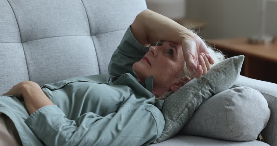 Sad unhappy senior old woman lying on back, resting on home sofa, feeling bad, sick, suffering from headache, depression, illness, loneliness, apathy, going, through grief, loss