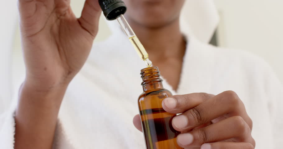 Happy biracial woman wearing bathrobe and holding bottle of oil in bathroom, slow motion. Domestic life, wellbeing and selfcare, unaltered.