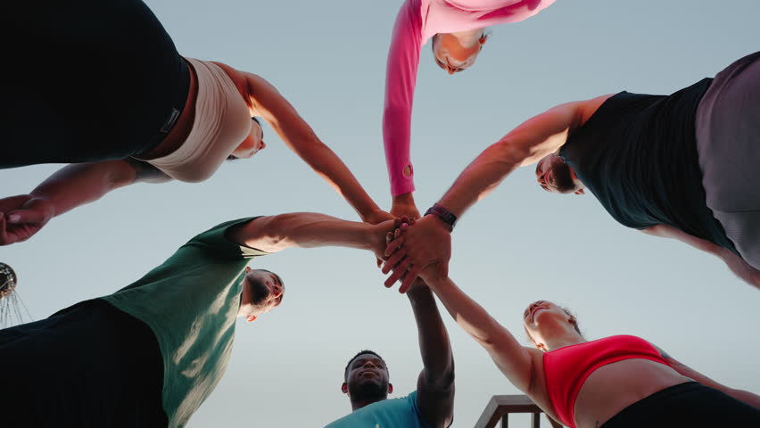 Sportive people make gesture of unity standing in circle. Professional athletes show solidarity in pursuit of healthy lifestyle standing under clear sky slow motion low angle shot