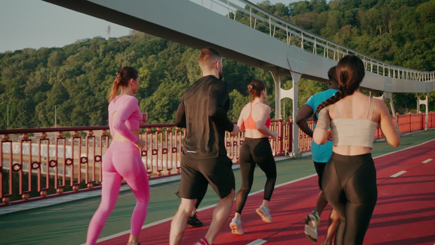 Athletes run on red running track on footbridge over river. Healthy young people exercise body moving actively under rays of morning sun slow motion - Powered by Shutterstock - Get 15% off with code: PIKWIZARD15