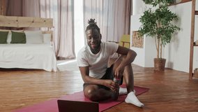 Young Beginner African American drinking bottle of water after workout, fitness, gymnastic, aerobic session. Muscular man talking to coach on videocall using laptop, smiles and laughs sitting on the