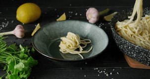 Person serving pasta with white sauce, putting spaghetti on plate using fork and spoon from pan, wooden table decorated with herbs, garlic, 4k footage, high quality horizontal video clip