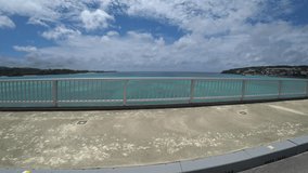 OKINAWA, JAPAN - JUL 2021 : Driving around Kouri Island bridge in sunny summer day. Wide camera, point of view (POV), seaside road driving. Japanese holiday, vacation and resort concept 4K video.