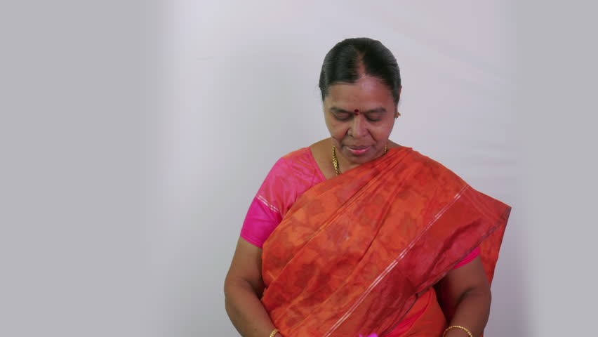 A South Indian woman in a traditional sari holds up a toy house in front of the camera. The background is plain white. The woman is smiling and looks happy.