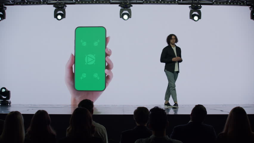 A man presenter is standing on a well-lit stage in front of a large LED screen showing a hand holding a generic smartphone with green screen. Technological, application or product launch presentation  Royalty-Free Stock Footage #1105692061