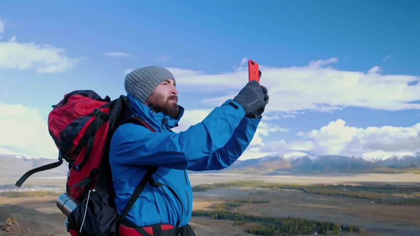 Hiker man taking picture with smart phone from mountain peak Strong man hiker taking photo with smart phone at mountain peak. Caucasian male walks with sticks and poses for camera on background of