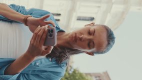 Vertical video, Beautiful girl with long brown hair, dressed in blue shirt, uses map application on her mobile phone in old town square