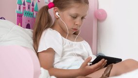 A little schoolgirl is watching a learning video on her phone.
The concept of a new distance education. The child enthusiastically looks at the phone while lying on the bed. 