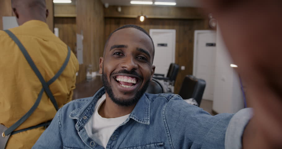 Joyful African American man taking selfie posing in barbershop smiling looking at camera while barber walking in background. Beauty and photography concept. Royalty-Free Stock Footage #1105721975