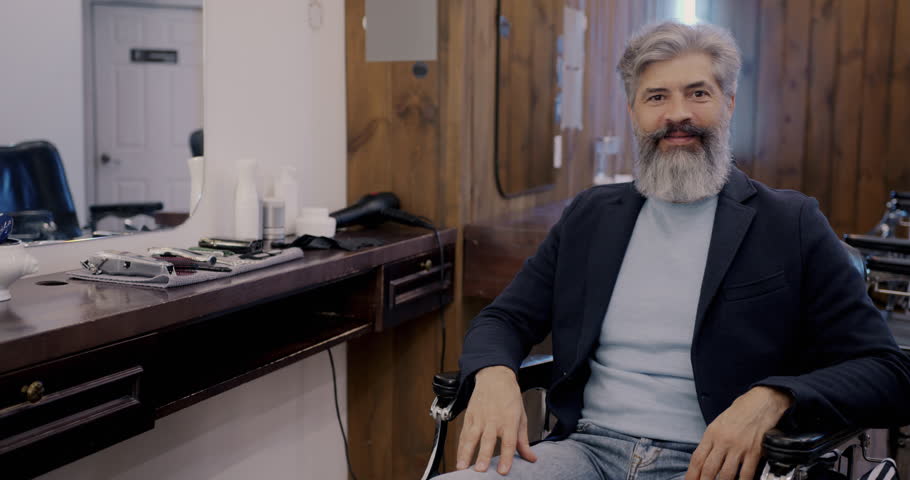 Slow motion potrait of smiling mature man sitting in armchair in barbershop and looking at camera. Style and masculine beauty concept.