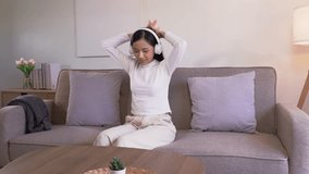 Cute Asian woman wear earphones listening music on smartphone sit on the sofa listen to music happy and relax at home