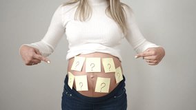 Young pregnant woman pointing to question marks reminder papers on belly asking for baby gender over isolated white background