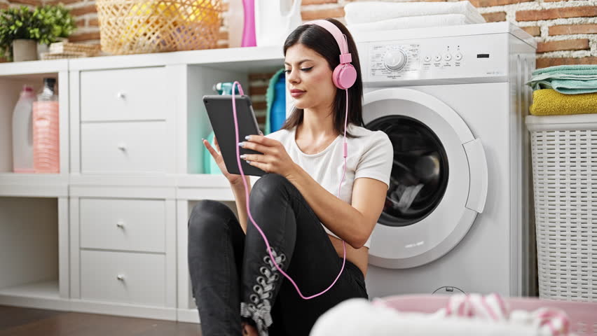 Young caucasian woman using touchpad and headphones waiting for washing machine at laundry room