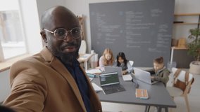 Medium shot of male African American teacher holding smartphone making vertical video of himself and elementary age multiracial students sitting at desks working with laptops