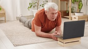 Mature senior man doing fitness exercises, gymnastics at home. Online lesson with laptop pc. Talking trainer. Recreation, well being. Elderly male exercising training, stretching. Old man working out.