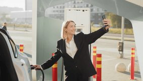 Cheerful caucasian woman in business suit having video call on modern smartphone while charging car at station. Busy modern lifestyles and innovation concept. Woman having video call on mobile.