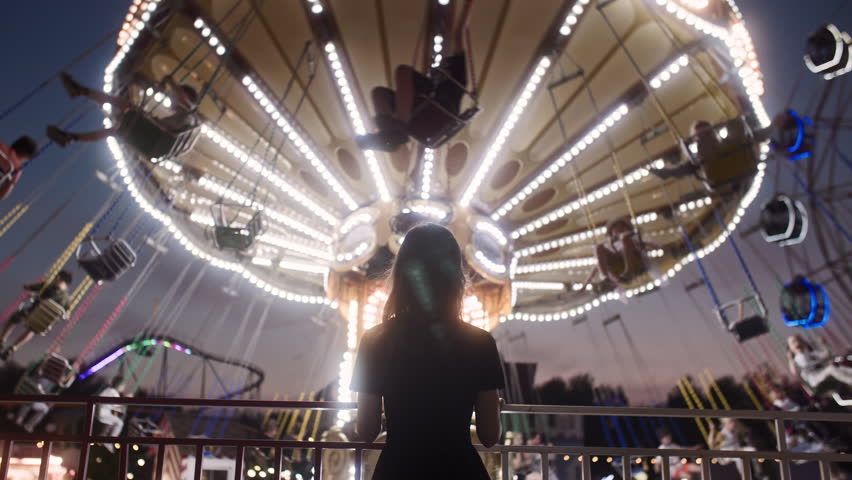 Rear view of an anonymous woman in dress standing by lighted carousel. In the background, people having fun riding on carousel in amusement park. Royalty-Free Stock Footage #1105746257