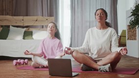 Young hispanic mom and cute little kid daughter meditating in lotus pose at home, happy healthy family mother and small child girl having fun doing yoga exercise relaxing together in living room.