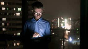 A man in a suit at work reads the news on a tablet. Outside the window the glowing windows of business centers. Overtime in the office, left alone at work. Outside the window a glowing night city - Powered by Shutterstock - Get 15% off with code: PIKWIZARD15