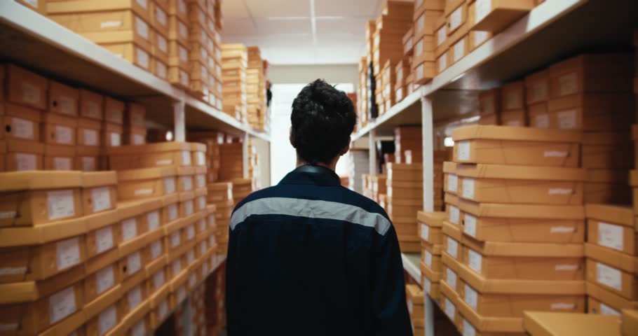 Rear view of a man in a blue uniform walking through a warehouse with yellow boxes in a shoe store. Video filmed in high quality Royalty-Free Stock Footage #1105750057