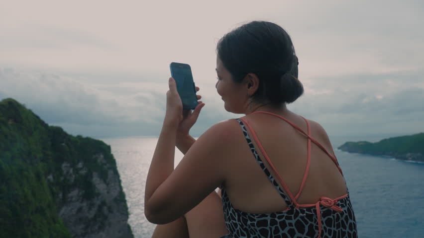 Asian girl relaxing on a rock above sea in summer and recording video on cell phone. Japanese woman holding hand phone sitting on stone at edge of cliff on ocean. Female online social media. Evening 