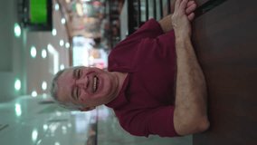 Happy senior man laughing and smiling sitting inside Brazilian cafeteria. Joyful caucasian elderly person in restaurant business in vertical video