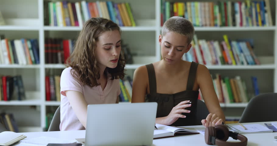 Two serious teenage student girls collaborating on class project together, talking at laptop in library, writing notes, discussing research study. Successful student helping friend with homework task Royalty-Free Stock Footage #1105763521